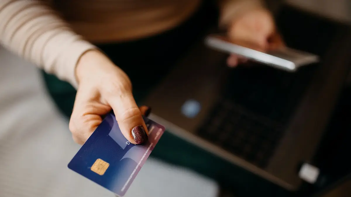 Mano de mujer que muestra una tarjeta de crédito azul con chip, con una notebook desenfocada detrás