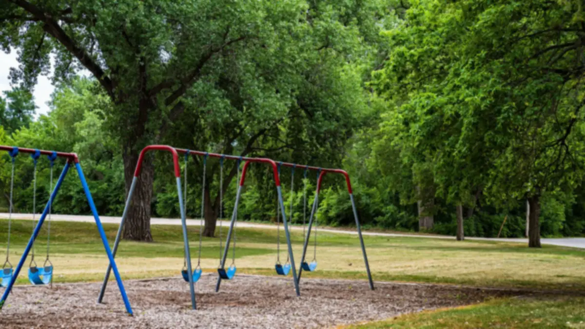 Hamacas vacías de una plaza, con asientos celestes y árboles frondosos detrás