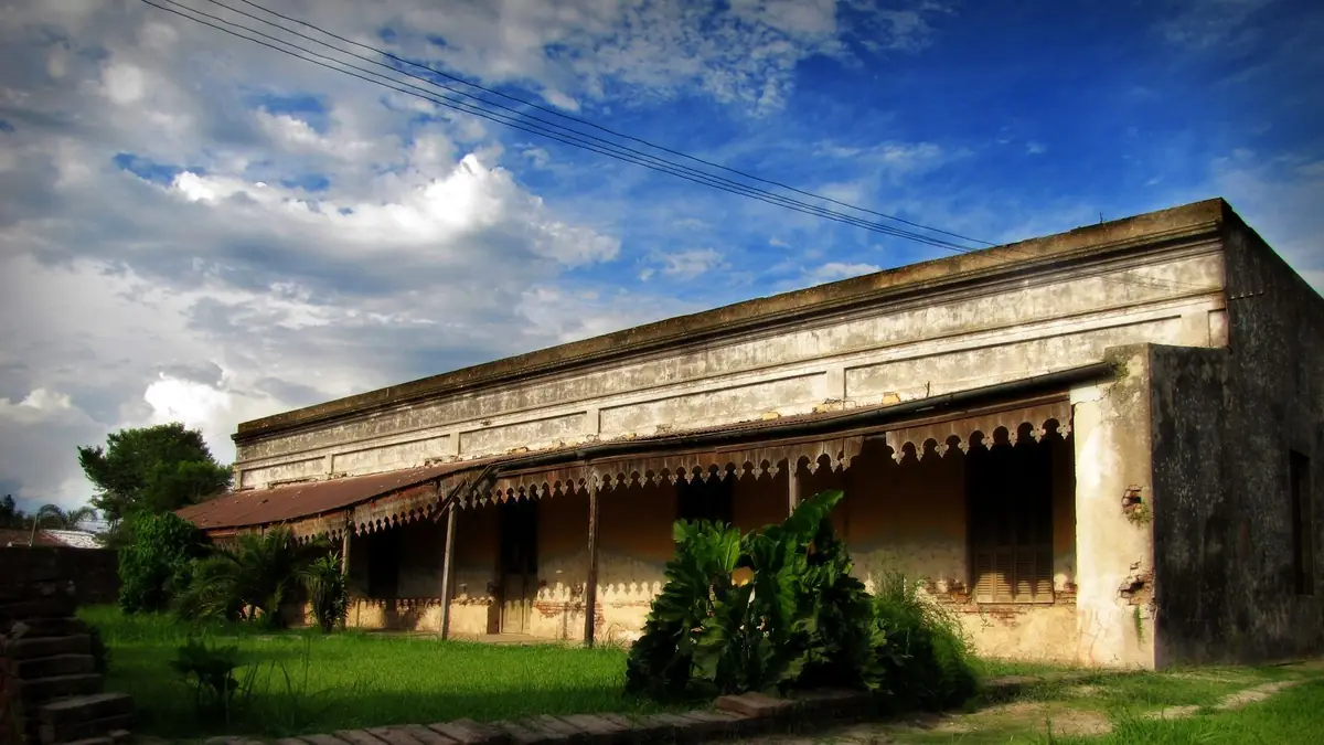 Casona antigua de una planta, con galería de chapa y paredes descascaradas, rodeada de pasto crecido bajo un cielo con nubes